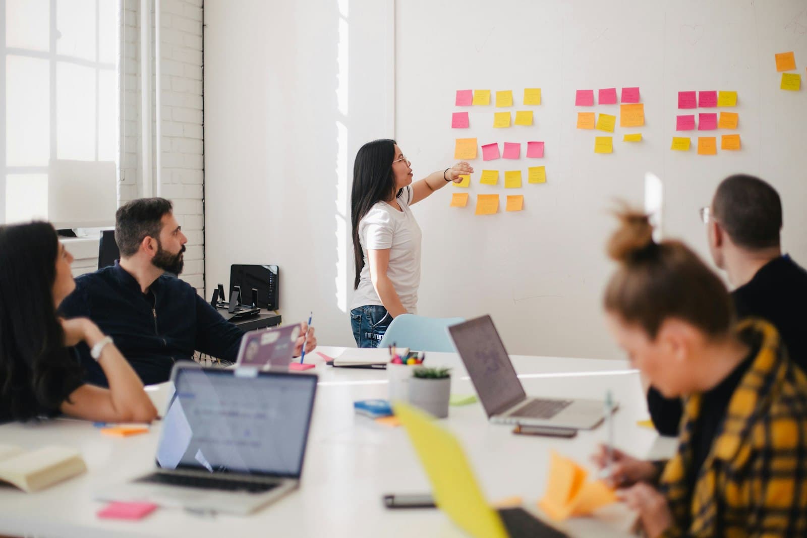 Team reviewing a kanban-style project board of sticky notes around a conference table with laptops — one workspace carrying leads through to delivery
