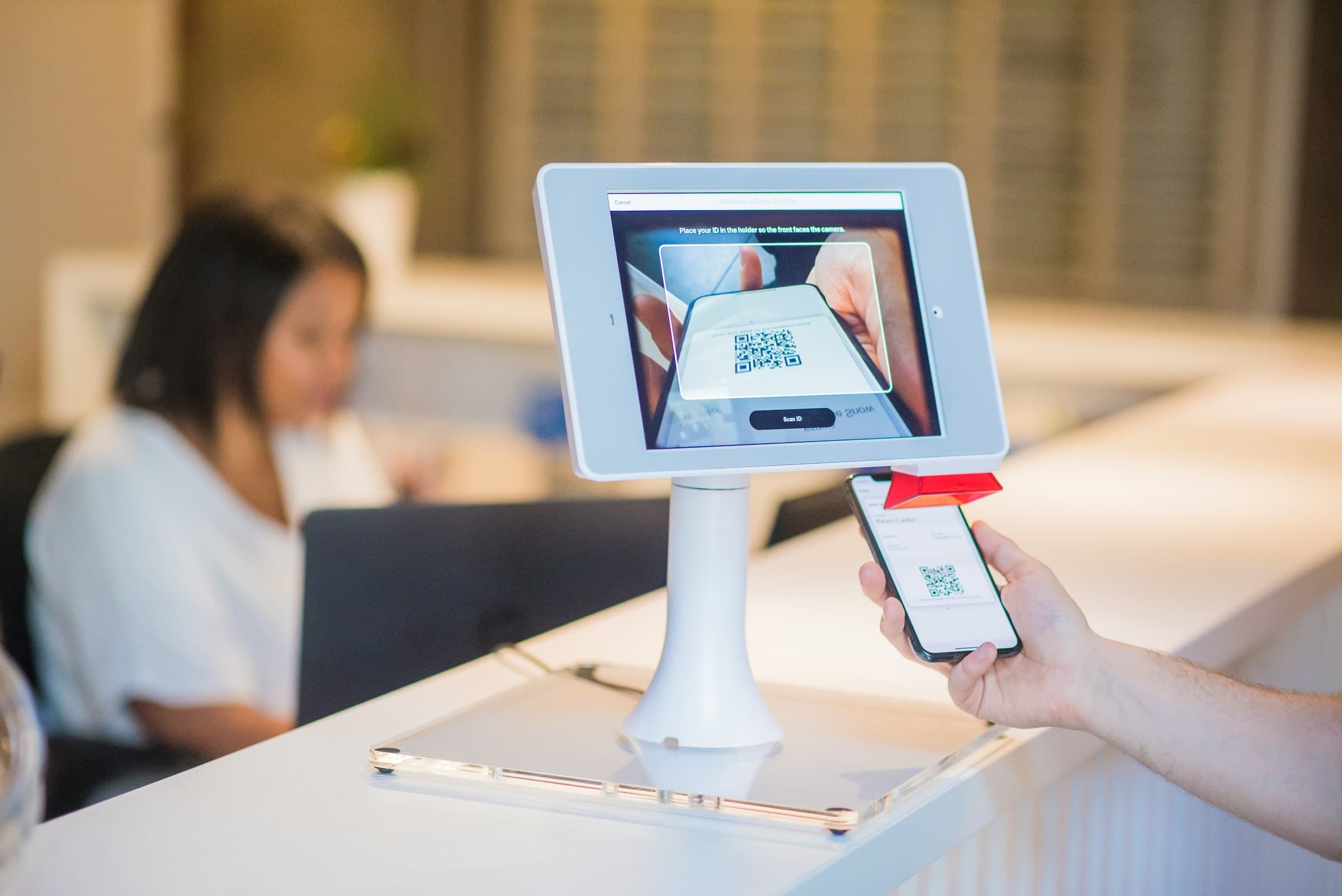 Visitor check-in tablet kiosk on a reception desk with QR scan and a receptionist in the lobby