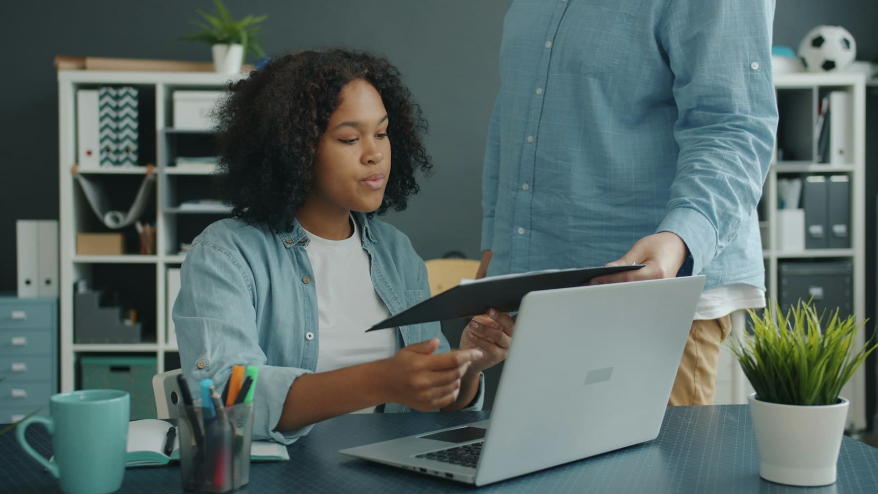 Two colleagues at a laptop, one handing over a folder of documents for review